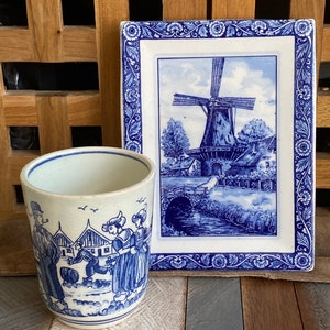 May include: A blue and white ceramic mug featuring a windmill scene and people in traditional Dutch clothing. The mug is beside a blue and white ceramic tile with a windmill scene.