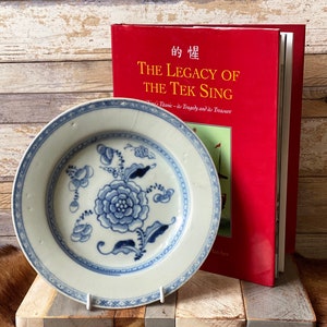 May include: A white porcelain plate with a blue and white floral design. The plate is sitting on a wooden surface next to a red book titled "The Legacy of the Tek Sing: China's Titanic - its Tragedy and its Treasure".