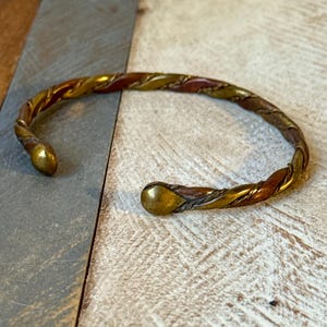 May include: A twisted metal cuff bracelet in gold, bronze, and copper tones. The open bracelet has rounded tips. The metal is intricately woven, creating a textured appearance. The bracelet is displayed on a light-coloured wooden surface.