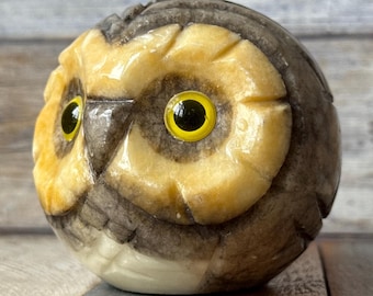 May include: A carved owl figurine with a round body. The owl has large yellow eyes, a light tan face, and a dark brown head. The owl's beak is not visible. The figurine is made of a stone-like material.