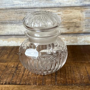 May include: A clear glass jar with a textured, patterned design. The jar has a lid with a similar pattern.