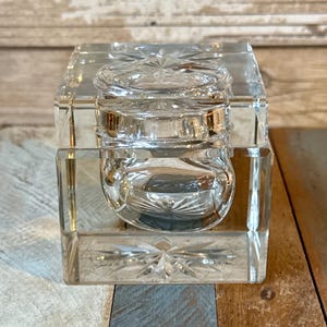 Può includere: Un calamaio di cristallo trasparente di forma quadrata con un calamaio rotondo e con coperchio all'interno. Il calamaio e il calamaio presentano disegni a stella sulla parte superiore e inferiore. Il calamaio è appoggiato su una superficie di legno.