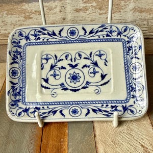 May include: Rectangular ceramic soap dish with a white base and blue floral design. The dish features a detailed border and central floral motifs. The design is reminiscent of classic blue and white porcelain. The dish is supported by a white metal stand.