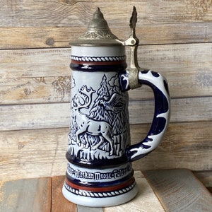 Pu&ograve; includere: Una boccale di birra in ceramica bianca con un design blu e bianco di un alce, un uccello e alberi. La boccale ha un coperchio in metallo con un manico e un manico blu e bianco. La boccale ha le parole "Alaskan Moose Falcon" sul fondo.