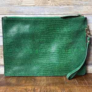 Può includere: Una pochette verde con una texture simile a quella di un rettile. La borsa rettangolare ha una chiusura a cerniera e un cinturino da polso attaccato con un fermaglio color oro. Lo sfondo è una superficie di legno.