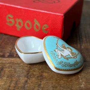 Puede incluir: Una caja de porcelana en forma de corazón con un diseño azul y dorado. La tapa presenta un querubín en un columpio con detalles dorados. La caja está hecha por Spode.