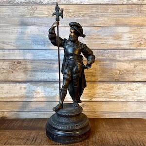 May include: A bronze statue of a man in historical attire, holding a halberd and a sword. The figure is standing on a round base. The statue is dark brown with bronze highlights, and the background is a wooden wall.