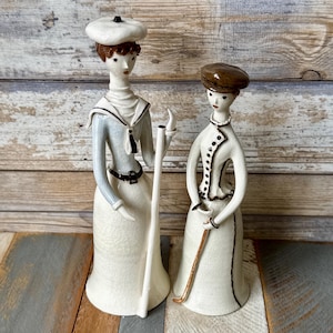 May include: Two ceramic figurines depicting figures in vintage attire. The taller figure wears a white beret, a light blue sailor-style top, and a long cream skirt. The shorter figure wears a brown hat, a cream jacket with dark buttons, and a long cream skirt.