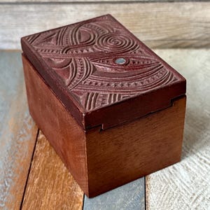 May include: A rectangular wooden box with a carved lid. The lid is a dark reddish-brown color and features intricate, swirling patterns. The box itself is a lighter brown, with a smooth finish. A small, round, silver-colored accent is set into the lid.