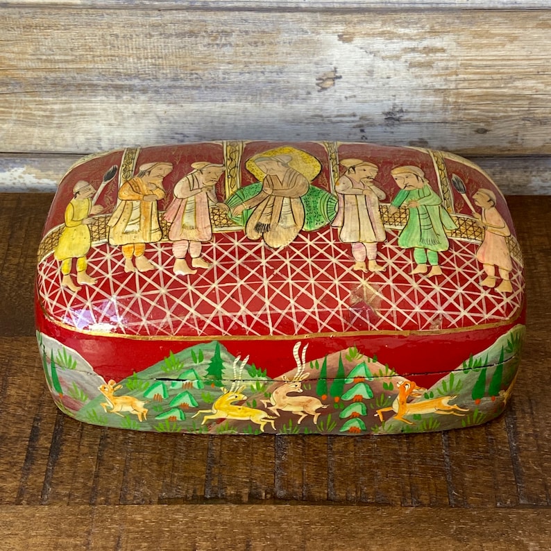 Op de afbeelding: Een rechthoekige houten doos met een rode basis en een gouden deksel. Het deksel is versierd met een handgeschilderd tafereel van mensen in traditionele kleding, en de basis is versierd met een handgeschilderd tafereel van herten in een bos.