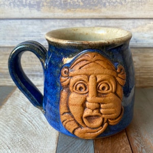 May include: A blue ceramic mug featuring a brown, textured face of a person with a surprised expression. The mug has a handle and is glazed with a blue and brown glaze.
