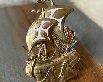 May include: A gold-toned metal brooch in the shape of a ship with white and red accents. The ship has a sail with a white cross design and blue accents on the hull.