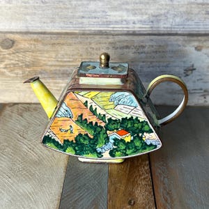 Könnte beinhalten: Eine kleine, gelbe, grüne und braune Cloisonné-Teekanne mit einer Landschaftsszene. Die Teekanne hat einen quadratischen Korpus mit einem abgerundeten Ausguss und Henkel.