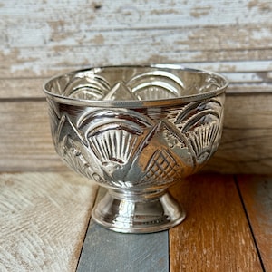 Puede incluir: Un cuenco plateado con pie y diseños en relieve intrincados. El cuenco presenta un patrón detallado de formas y texturas estilizadas, con una base de pedestal. La superficie del cuenco refleja la luz, resaltando sus elementos decorativos.