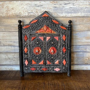 Puede incluir: Un espejo con marco de madera negro con intrincados detalles en plata y rojo. El espejo tiene dos puertas y está decorado con un patrón geométrico.