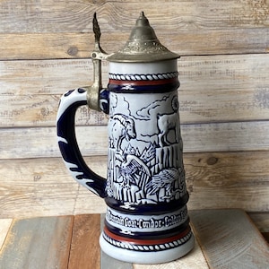 Pu&ograve; includere: Una boccale di birra in ceramica bianca con un design blu e bianco che presenta una capra di montagna, un condor e un'aquila reale. La boccale ha un coperchio in metallo con un manico.