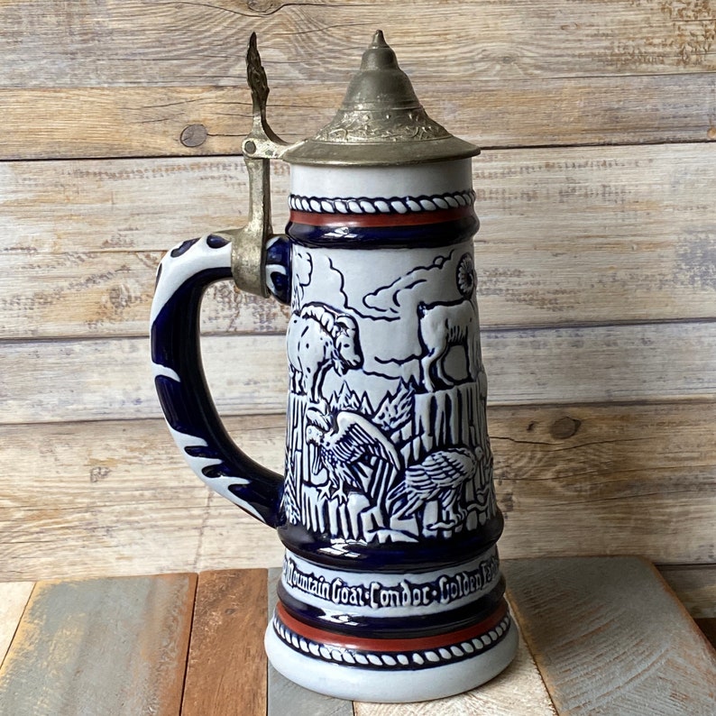 Pu&ograve; includere: Una boccale di birra in ceramica bianca con un design blu e bianco che presenta una capra di montagna, un condor e un'aquila reale. La boccale ha un coperchio in metallo con un manico.