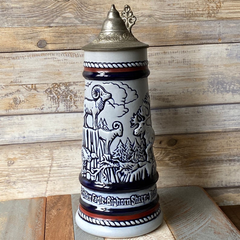 Pu&ograve; includere: Una boccale di birra in ceramica bianca con un coperchio argentato e illustrazioni blu e bianche di animali, tra cui un ariete, un alce e un uccello. Il boccale ha una fascia blu con il testo "Golden Eagle, Bighorn Sheep, Moose" intorno alla base.
