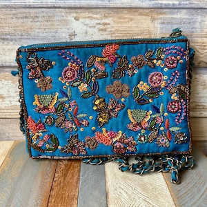 May include: Teal-colored clutch with intricate floral embroidery in shades of orange, pink, and green. The bag features a silver chain strap and a zipper closure. The floral design includes beads and sequins, adding texture and sparkle to the accessory.