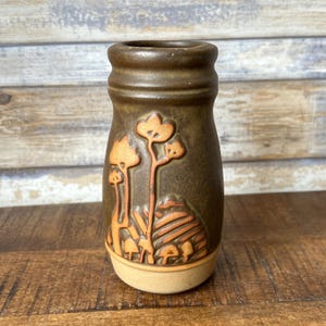 May include: A small, brown ceramic vase with a raised design of trees and mushrooms. The vase has a light brown rim and base.