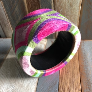 Könnte beinhalten: Ein farbenfrohes Armreif-Armband mit einem geometrischen Muster in Rosa-, Grün- und Lilatönen. Das Armband ist aus Stoff und hat ein schwarzes Innenleben.