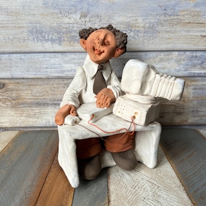 Pode incluir: Uma figurinha de cerâmica de uma pessoa sentada em uma mesa com um computador. A pessoa está sorrindo e tem cabelo castanho. O computador é branco e tem um monitor, teclado e mouse. A figurinha é pintada em tons de marrom, branco e laranja.