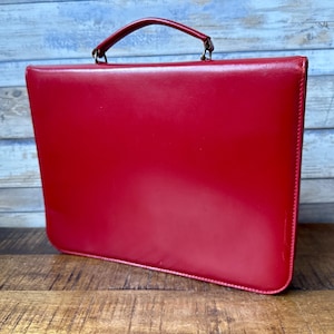 Puede incluir: Un maletín de cuero rojo con asa superior y herrajes metálicos. El maletín rectangular tiene esquinas redondeadas y está sobre una superficie de madera. El fondo es de madera pintada de azul y blanco desgastado.