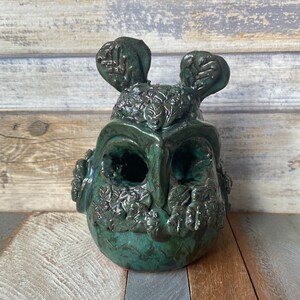 Puede incluir: Una escultura de cerámica verde de una cara con detalles de hojas. La escultura tiene dos orejas grandes y una superficie texturizada.
