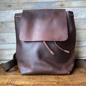 Puede incluir: Una mochila de cuero marrón con cierre de solapa y cordón de ajuste. La mochila tiene un diseño sencillo y está hecha de cuero de alta calidad.
