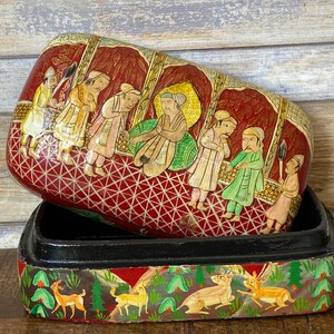Op de afbeelding: Een rood en goud geschilderde houten doos met een scharnierend deksel. Het deksel toont een sc&egrave;ne met mensen in traditionele kleding, terwijl de bodem van de doos een sc&egrave;ne met herten in een bos toont.