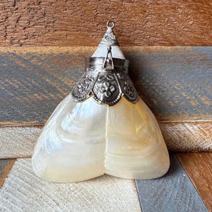 Könnte beinhalten: Perlmuttfarbene, muschelförmige Parfümflasche mit silberfarbenem Metallaufsatz und dekorativem Filigran. Die Flasche hat eine konische Spitze und eine Schlaufe zum Aufhängen. Die Muschel hat einen cremeweißen und gelben Farbton.