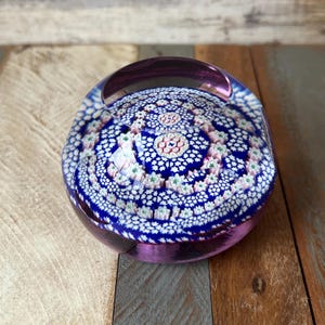 May include: A purple glass paperweight with a white and blue patterned design. The pattern is made up of small circles and stars.