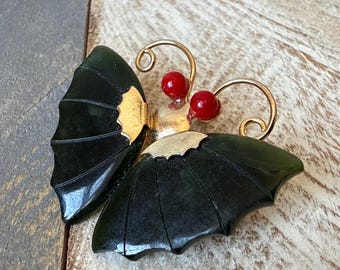 May include: A butterfly brooch with dark green jade wings and gold-coloured accents. The brooch features two red beads for the eyes and delicate gold-coloured antennae. The wings have a detailed, layered design.