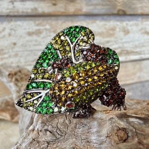 Puede incluir: Un broche con forma de rana sobre una hoja, cubierto de pequeños cristales brillantes en tonos verde esmeralda, lima, amarillo y marrón oscuro. El engaste de metal es oscuro, lo que crea un contraste con las piedras multicolores.