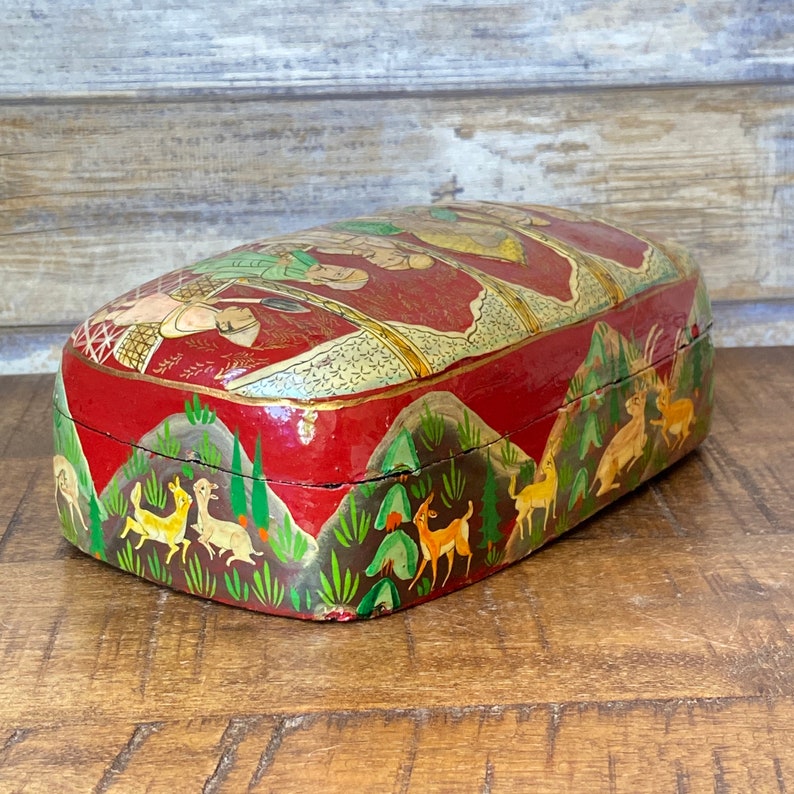 Op de afbeelding: Een rode houten doos met een scharnierend deksel, versierd met handgeschilderde sc&egrave;nes van mensen en dieren. De doos is in een traditionele stijl geschilderd met gouden accenten.