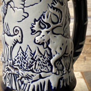 Pu&ograve; includere: Una boccale di birra in ceramica bianca con un design blu e bianco di un alce, un ariete e alberi. La boccale ha un manico e una base blu.