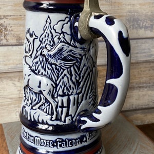 Pu&ograve; includere: Una boccale di birra in ceramica bianca con un design blu e bianco di un alce, un falco e montagne. La boccale ha un coperchio in peltro e un manico blu e bianco.