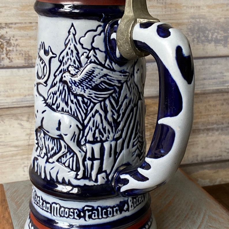 Pu&ograve; includere: Una boccale di birra in ceramica bianca con un design blu e bianco di un alce, un falco e montagne. La boccale ha un coperchio in peltro e un manico blu e bianco.