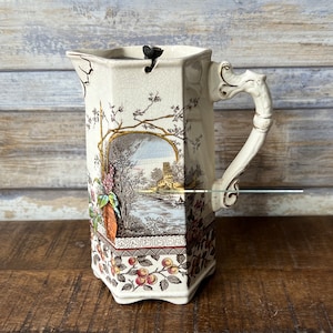 Può includere: Un'anfora in ceramica bianca con un motivo floreale marrone e dorato. L'anfora ha un coperchio e un manico. Il disegno presenta una scena paesaggistica con un castello e un fiume.