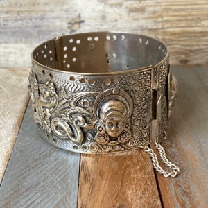 Könnte beinhalten: Silberfarbener Armreif mit einem komplizierten Design, das ein Frauen Gesicht und einen Drachen zeigt. Der Armreif hat eine Kette.