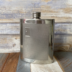 May include: A silver metal hip flask with a textured surface and a plain rectangular design on the front.