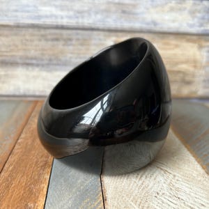May include: A glossy, black, oval-shaped decorative bowl. The bowl has a smooth, reflective surface and a slanted opening. The bowl is sitting on a wooden surface with various shades of brown, gray, and blue.