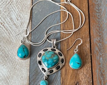 May include: A silver necklace and earrings set with turquoise and black stones. The necklace features a pendant with a large turquoise stone surrounded by smaller black stones. The earrings are teardrop shaped with turquoise stones.