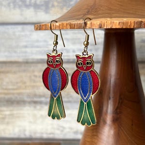 May include: A pair of dangle earrings featuring a red, blue, and green owl design. The owls have gold accents and are suspended from gold hooks.