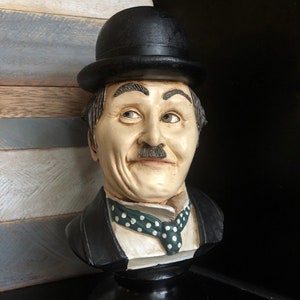 Pode incluir: Um busto de cerâmica de um homem usando um chapéu-coco preto e uma gravata estampada. O busto é pintado com cores realistas e tem uma expressão facial detalhada.