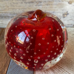 Può includere: Una mela di vetro rosso con un gambo di vetro trasparente e una superficie testurizzata. La mela è ricoperta da piccole bolle d'aria.