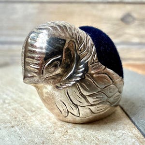 May include: A silver-toned metal pincushion shaped like a bird with engraved feather details and a dark navy blue velvet cushion insert. The bird figure is depicted in a resting position with its head tucked slightly forward and eyes closed.
