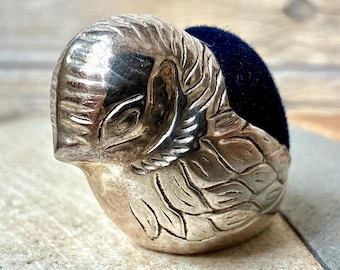 Op de afbeelding: Een zilverkleurig metalen speldenkussen in de vorm van een vogel met gegraveerde veertjes en een donker marineblauw fluwelen kussentje. De vogel is afgebeeld in ruststand met de kop licht naar voren gebogen en de ogen gesloten.