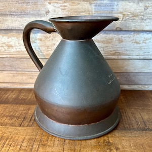 May include: A dark metallic pitcher with a wide, flared rim and a curved handle. The body has a rounded, conical shape with a flat base. The surface shows a matte, weathered finish with subtle copper-toned highlights near the bottom and rim.