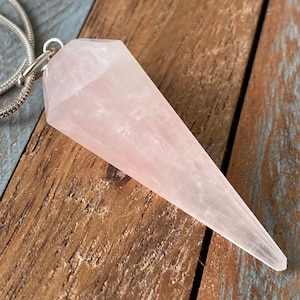 Op de afbeelding: Een lichtroze rozenkwartskristal hanger in de vorm van een punt, hangend aan een zilveren ketting.